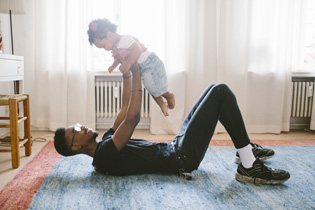 A father lieing down on a rug holding a toddler up in the air.