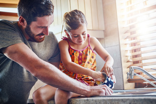 A dad helps his daughter wash a mug while sitting on the kitchen counter beside the sink.