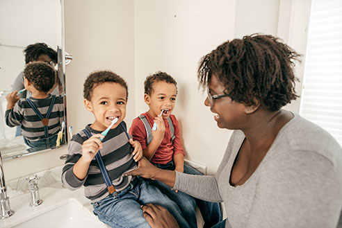 A grandmother helping two small children, sitting together on the bathroom counter, brushing their teeth.