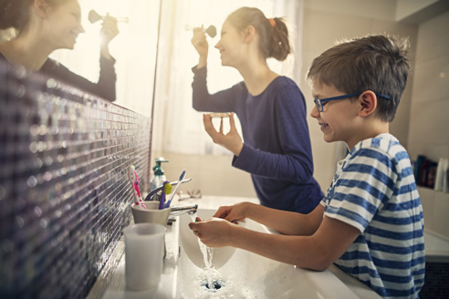 Parent applying makeup while child washes at same sink and mirror.
